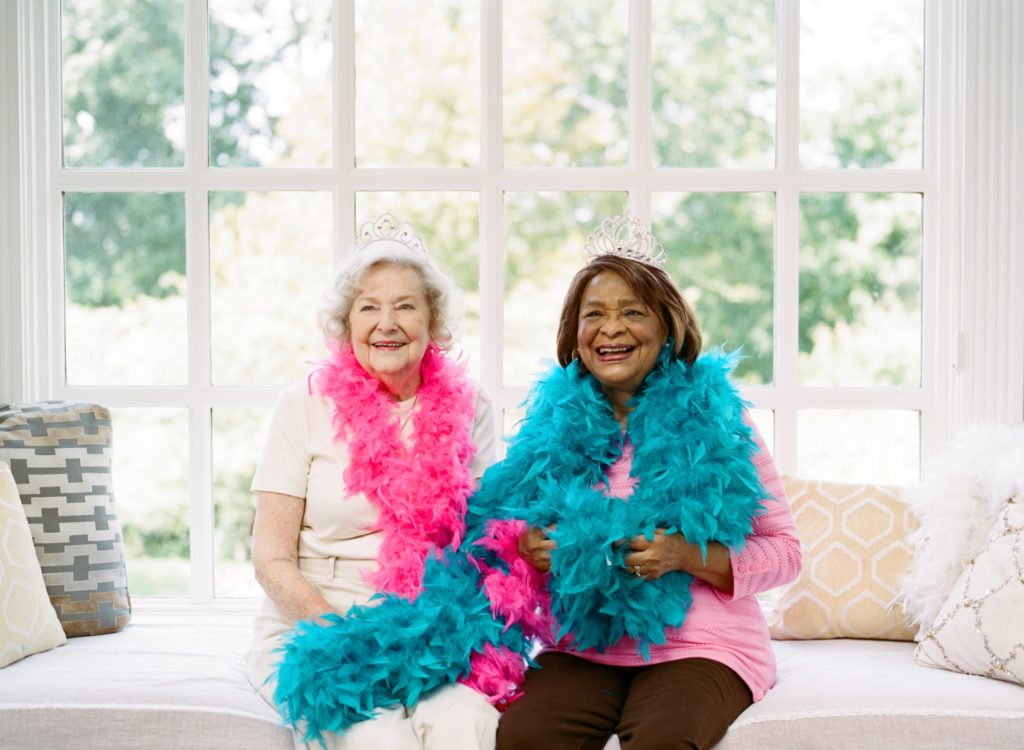 Senior woman with caregiver, both wearing feather boas around their necks seated on interior window bench | Comfort Keepers Calgary | BLOG POST | Osteoporosis Prevention