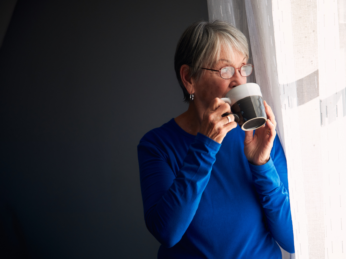 A senior woman drinking a cup of coffee looks out a window | Maintaining Your Loved One’s Well-Being During Self-Isolation in Edmonton | Comfort Keepers Edmonton