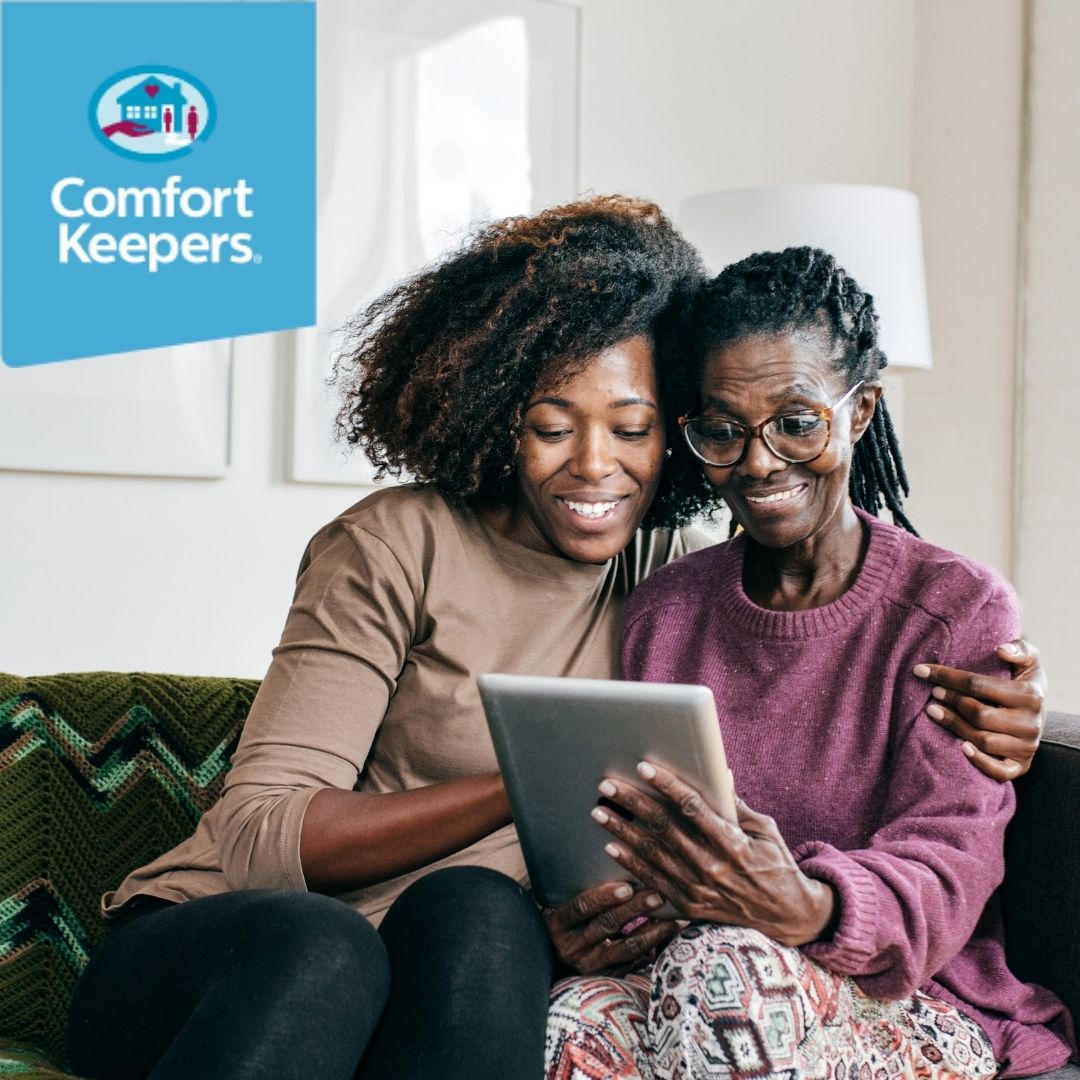 two women sitting on the couch. The younger women with her arm around the older. Both are smiling and looking at a tech device