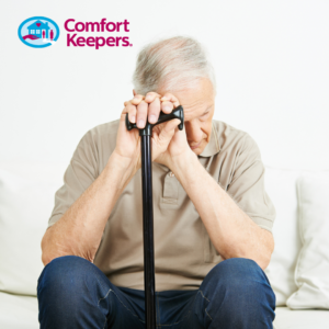 Post-Hoiday Blues Comfort Keepers logo featured in top left corner. Senior male seated on white couch holding a cane between his knees and his head is bent forward. Conveying depression.