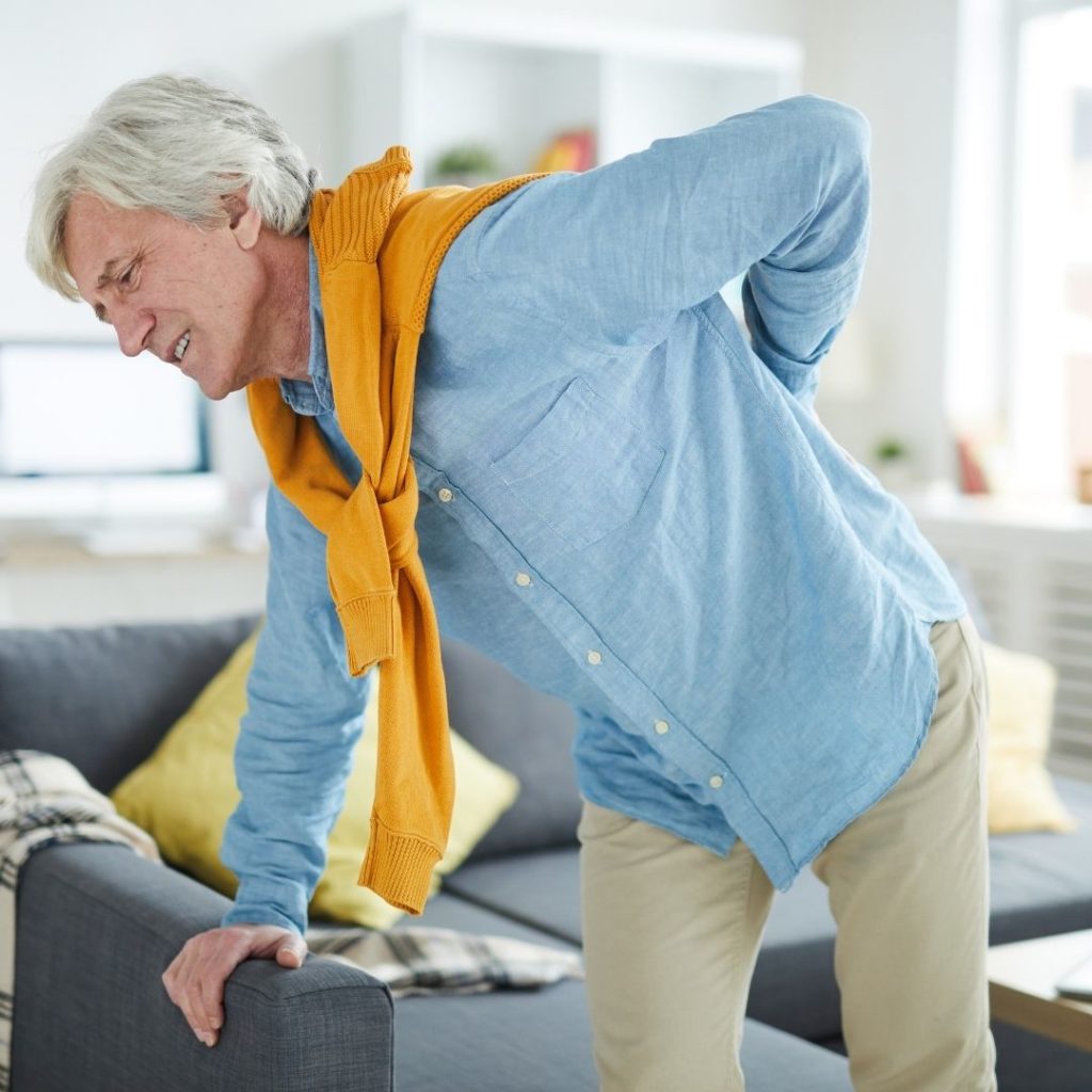 Comfort Keepers Edmonton - Blog: Seniors Living with Chronic Pain - Senior Adult leaning on grey couch for support while placing other hand on back seemingly in pain