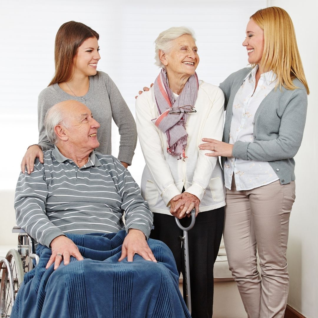 Group of four individual with arms around two senior citizens. One with a cane, the other in a wheelchair with blanket over lap