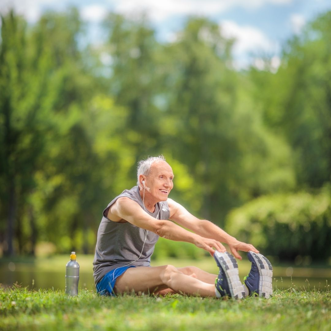 Senior citizen sitting on the grass and stretching legs. Next to him sits a bottle of water.