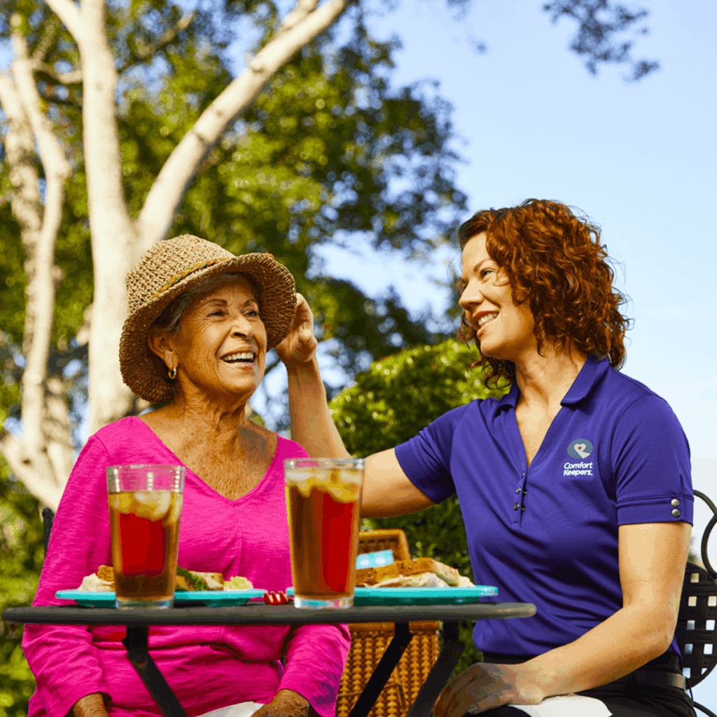This is a bright, outdoor photograph featuring two women, likely a senior client and a professional caregiver of Comfort Keepers Guelph, enjoying a meal together.