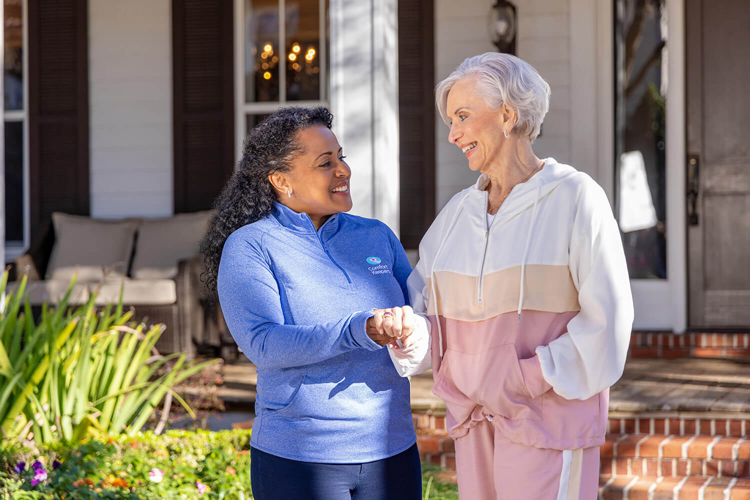 female caregiver with brown hair and wearing a blue uniform shirt sits next to an elderly woman with short, white hair on a couch. The caregiver is smiling and gently holding the senior woman's hand while looking at her, suggesting a supportive and comforting interaction. The senior woman is looking toward the camera.