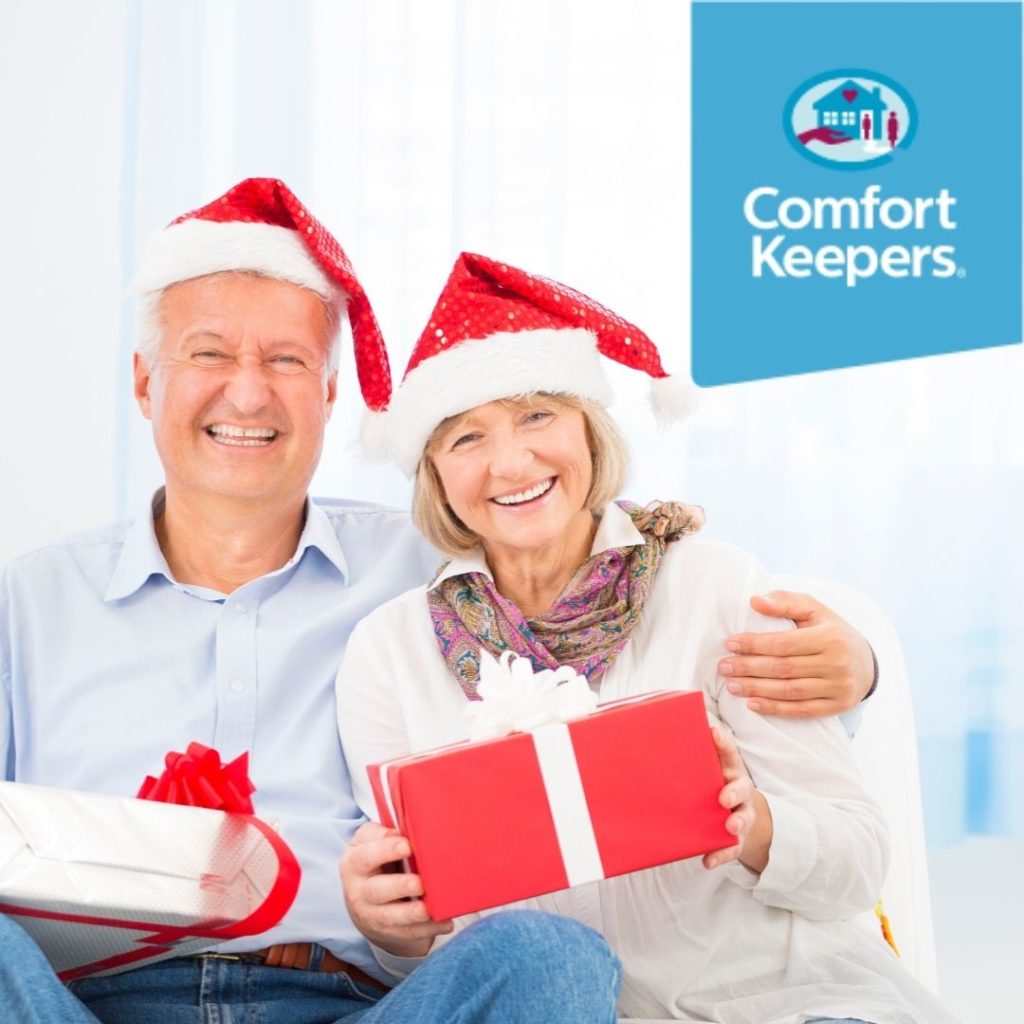 Two seniors wearing Santa hats sit embraced with gifts in hand | Comfort Keepers North and West Vancouver