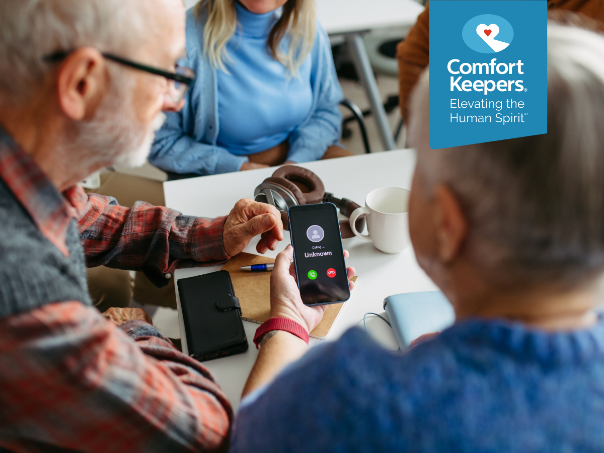 A pair of seniors seated at a table view an unknown caller coming through their mobile phone | Holiday Scams: What Seniors in BC Should Look Out For | Comfort Keepers North and West Vancouver