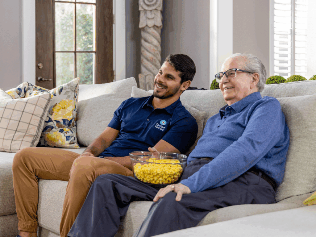 A senior male and caregiver enjoy a bowl of popcorn while seated on a couch | How to Know When a Loved One Needs Senior Care | Comfort Keepers North and West Vancouver | BLOG POST