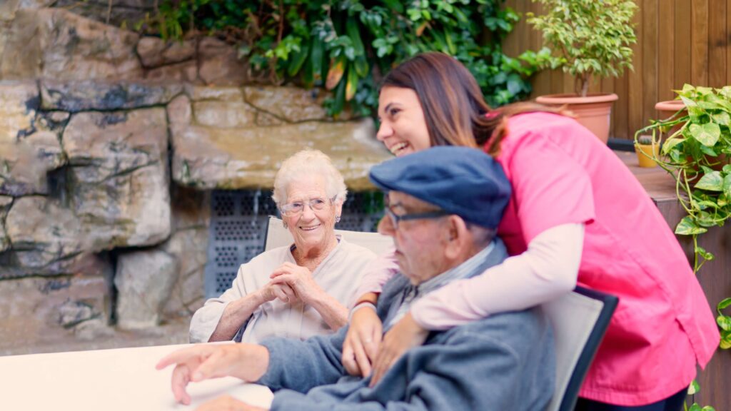 A caregiver sharing laughter with a group of seniors in a garden setting, emphasizing emotional support for Parkinson’s home care.