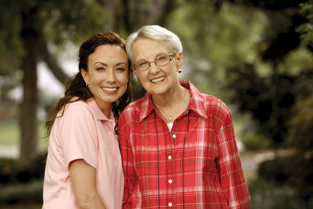 smiling, middle-aged female caregiver wearing a blue quarter-zip shirt with a Comfort Keepers logo, standing outside on a porch holding hands with a smiling elderly woman with short white hair. The senior woman is wearing a pink and white zip-up hoodie and matching pants. They are looking into each other's eyes, suggesting a warm and friendly connection.