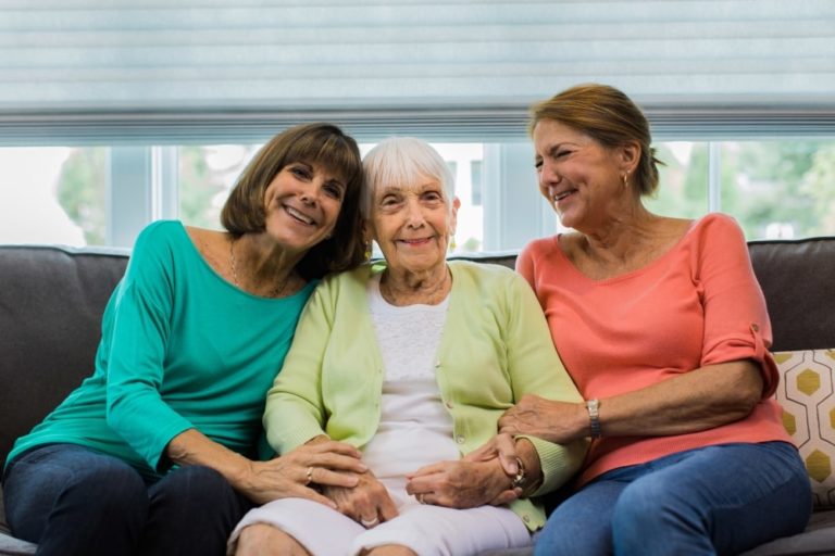 An elderly woman sits comfortably on the chair at home, surrounded by her two happy family members who are sitting one on either side.