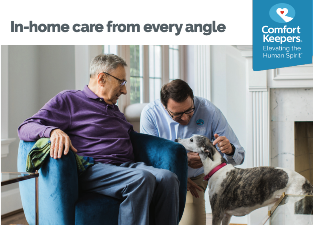 A Male senior, happy and comfortable sitting in his chair at home with the comfort keepers in home care assistant next to him holding his beloved dog. (With Comfort Keepers Logo on the upper right side and their mission "In-Home Care from every angle") 🐾