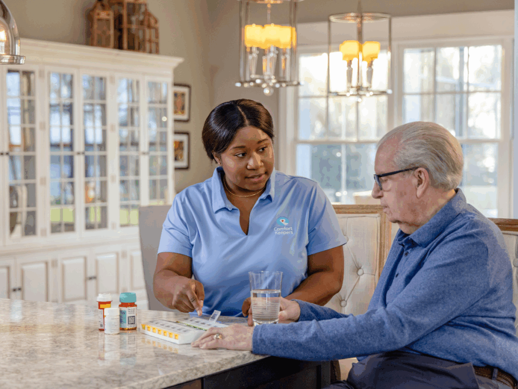 Senior male is seated in kitchen discussing medications with caregiver | Understanding the Differences Between Companionship, Personal Care, and Nursing Care at Home in Ridge Meadows | Comfort Keepers Ridge Meadows | BLOG POST