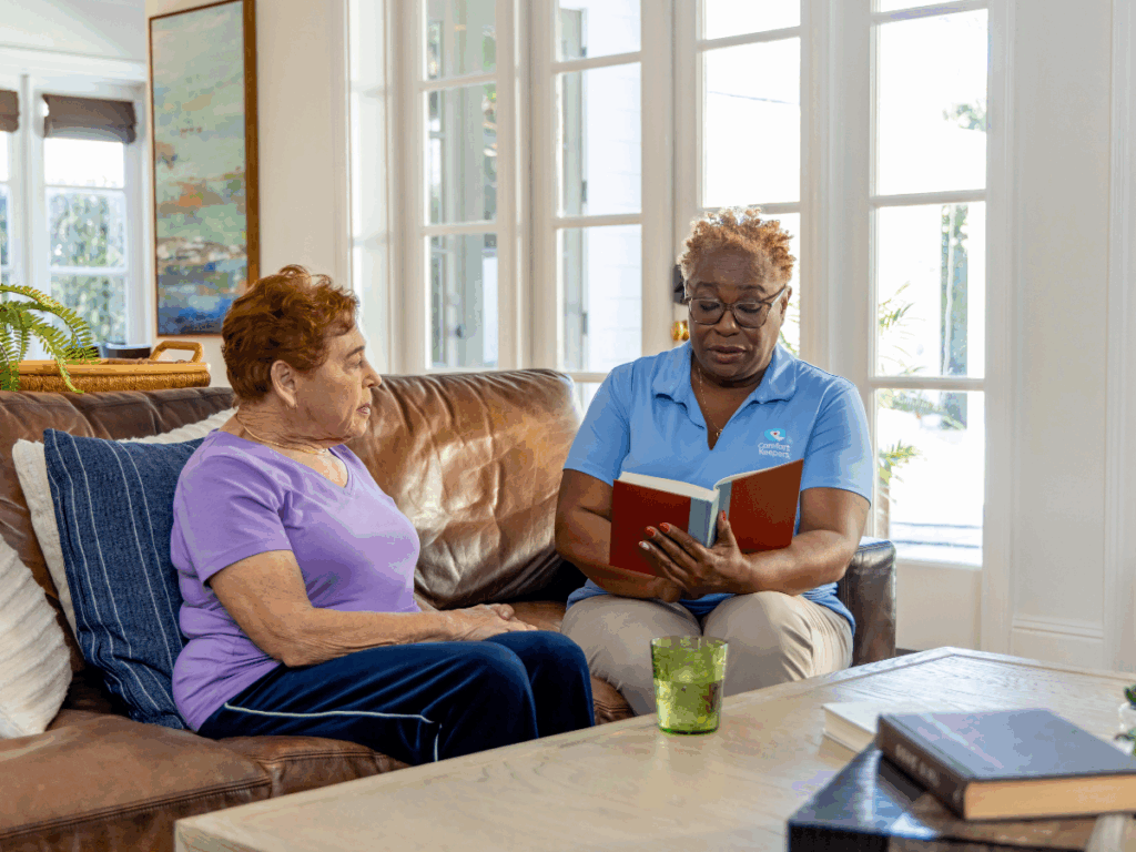 Senior woman seated on couch with caregiver who reads a book aloud | The Benefits of At-Home Dementia Care: A Comparative Guide for Tri-Cities Families | Comfort Keepers Tri-Cities