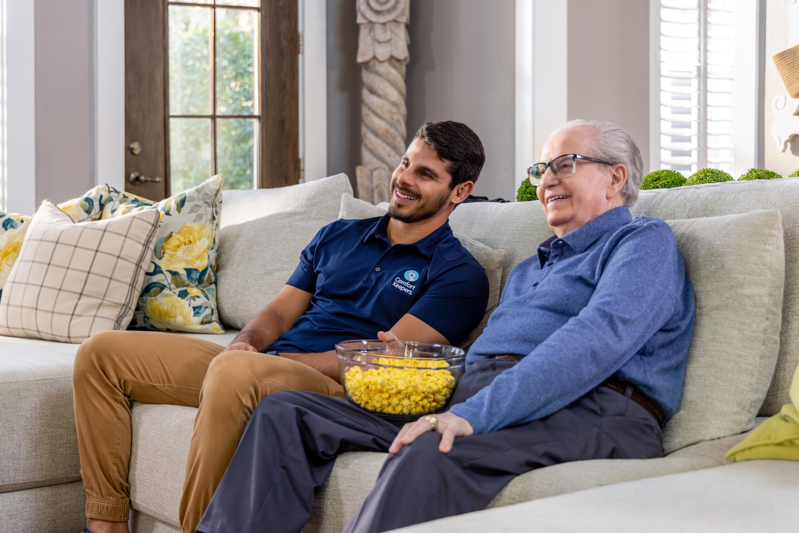 Senior male with popcorn seated on the couch next to caregiver | What is Interactive Caregiving? | Interactive Caregiving Tri-Cities | Comfort Keepers Coquitlam | BLOG POST