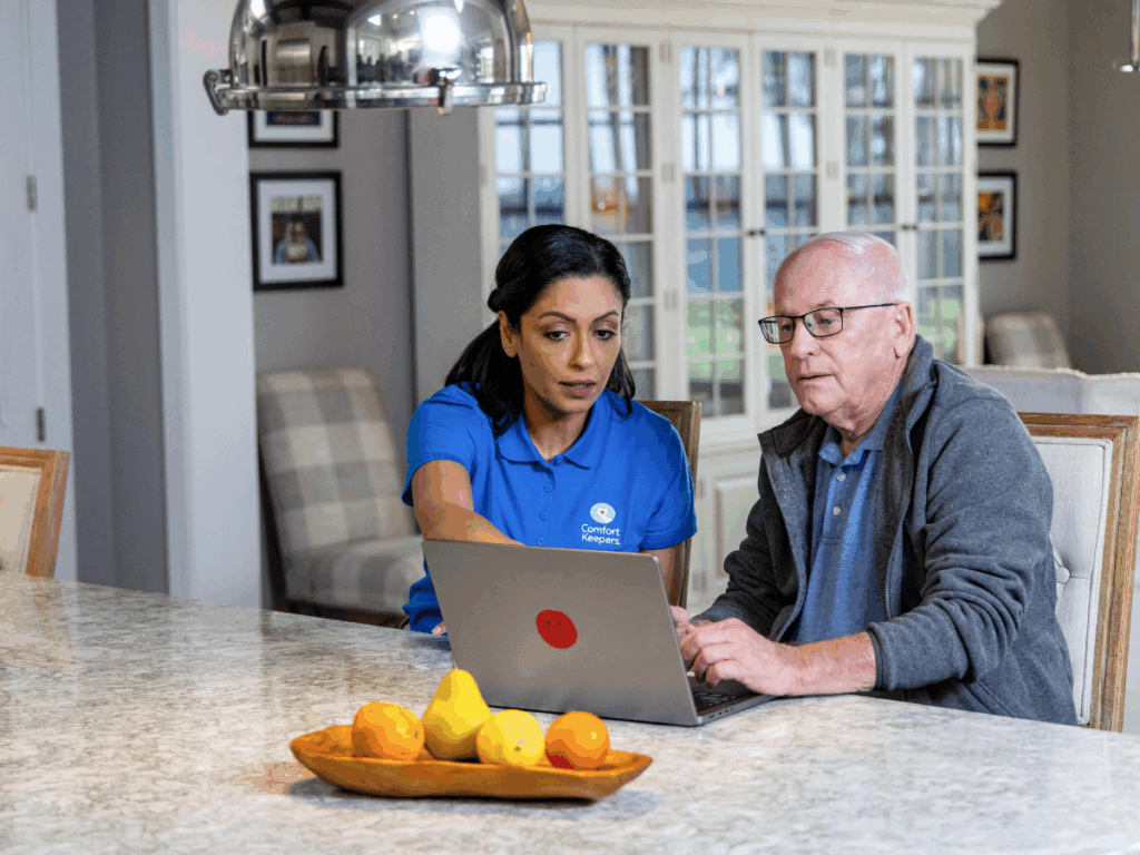 Senior male seated in kitchen reviewing computer with caregiver | The Complete Guide to Creating a Senior Home Emergency Plan in Victoria, BC | Comfort Keepers Victoria | BLOG POST